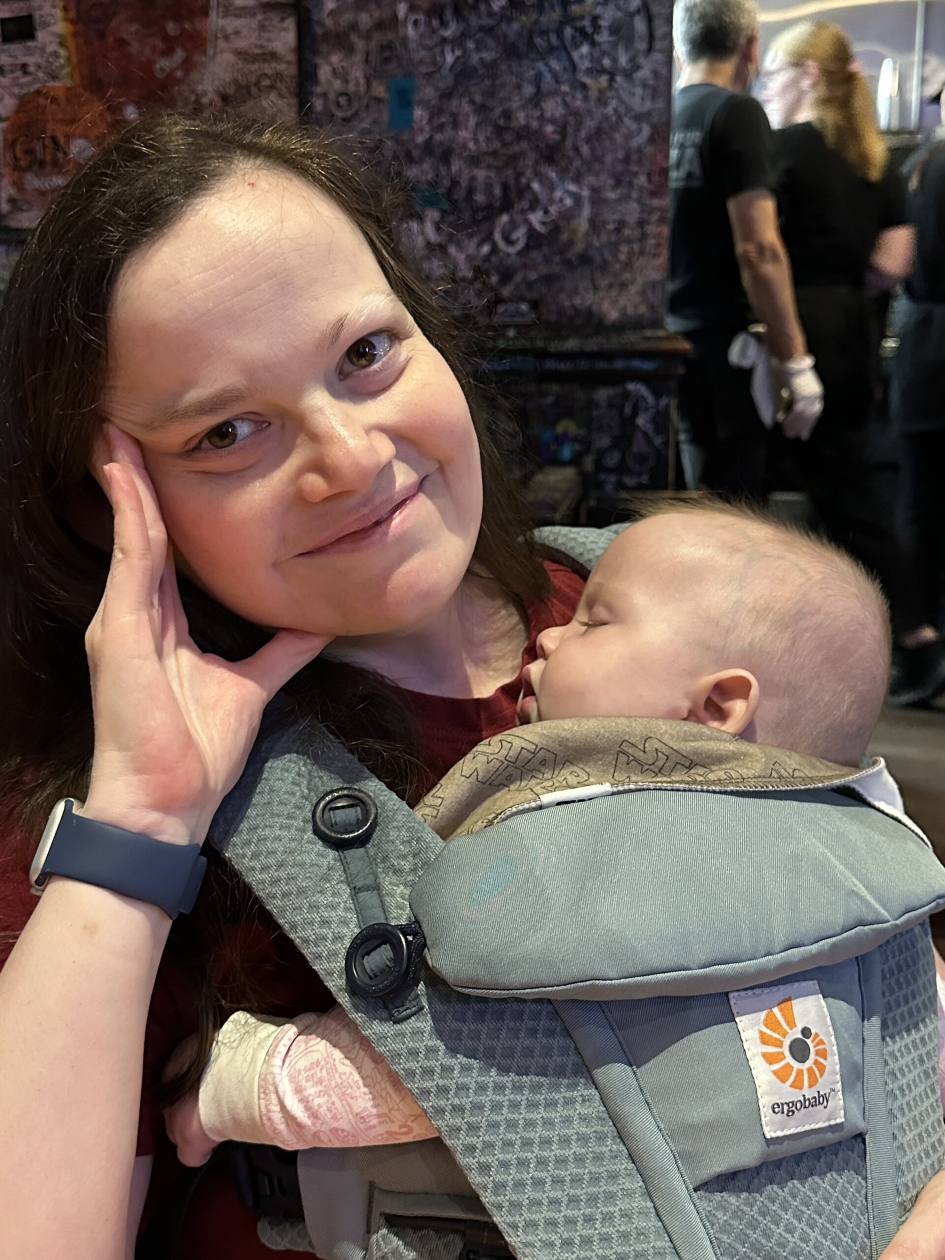 brunette woman smiling with baby sleeping in a body carrier